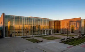 Modern building with glass facade reflecting sunset, featuring a sculpture and benches in the courtyard.