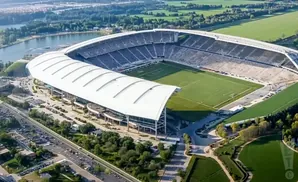 Aerial view of a large stadium with a white roof, surrounded by a football field, parking lots, and a body of water.