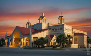 A large, tan building with red roofs and arched entrances stands under a colorful sunset sky.