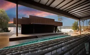 Outdoor amphitheater with tiered seating facing a stage, overlooking a lake at sunset.