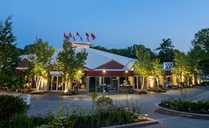 Exterior view of a building with a large white roof, red trim, and flags on top, illuminated at dusk.