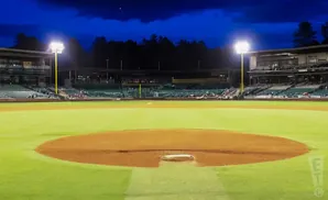 A baseball field at dusk with stadium lights on, and trees in the background.