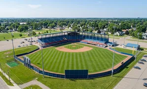 Aerial view of a baseball stadium with a manicured field, blue and red seating, and surrounding parking lots and trees.
