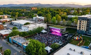 An outdoor concert with a crowd gathered in front of a stage, surrounded by buildings and trees, with mountains in the background.