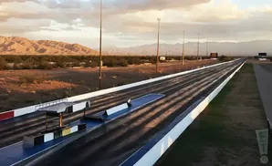 A drag strip stretches into the distance against a backdrop of desert mountains and a cloudy sky.