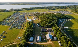 Aerial view of a large campground with many tents and cars next to a lake and forest.