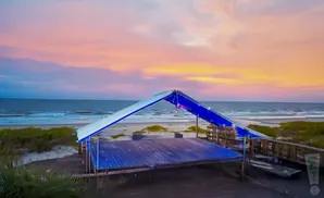 A wooden stage with a blue canopy sits on a beach at sunset.
