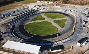 Aerial view of a race track with grandstands, a green infield, and surrounding buildings.