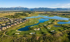 Aerial view of a golf course with lakes and homes, with mountains in the distance under a clear blue sky.