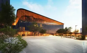 Modern glass building with curved roof and orange-lit interior, surrounded by trees and a paved plaza.