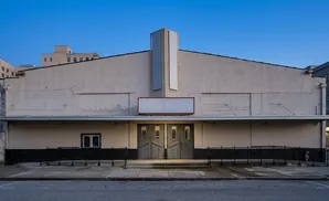 An old, beige movie theater with large double doors and a blank marquee sits under a clear blue sky.