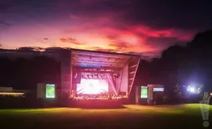 An outdoor stage with a bright screen and lighting is set up on a grassy field, with a vibrant sunset in the background.
