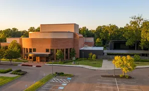 An aerial view of a modern brick building with an outdoor stage area, surrounded by trees during sunset.