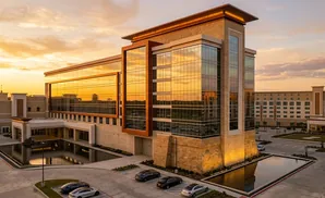 Modern hotel building with glass facade reflecting sunset, reflecting pool, and parked cars.