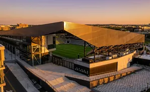 An aerial view of a modern soccer stadium at sunset with a large, angular roof and surrounding buildings.