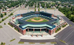 Aerial view of a baseball stadium with empty parking lots and surrounding residential areas and trees.
