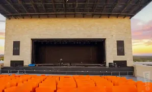 An outdoor amphitheater stage with orange seats in the foreground and a colorful sunset sky.
