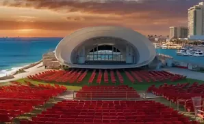 An outdoor amphitheater with red seats facing a shell-shaped stage by the ocean at sunset.