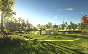 A golfer on a putting green with a pond and sand traps in the background under a sunny blue sky.