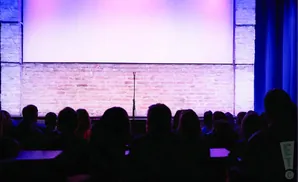 A comedy club stage with a brick wall backdrop, microphone, and audience silhouettes in purple light.