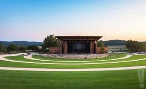An outdoor amphitheater with a stage and tiered grassy seating, surrounded by trees and rolling hills.