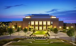 Modern brick building with large windows, illuminated at dusk, surrounded by manicured lawns and trees.