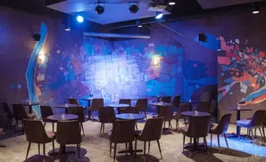 Dark room with colorful abstract murals, tables, and chairs, lit by blue spotlights.
