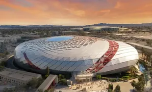 Aerial view of a modern, circular stadium with a patterned, white roof and red accents, set against a sunset sky.
