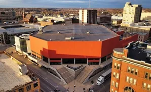 An aerial view of a modern, orange-walled building with tiered concrete stairs leading to its entrance, set against a city skyline.