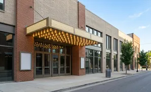 A theater marquee with bright lights illuminates the entrance of a brick building with large glass doors and windows.