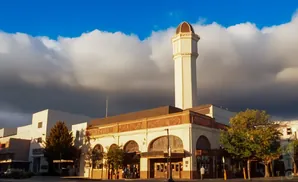A pale tower with a domed top rises above a tan building with arched windows, under a blue sky with white and dark clouds.