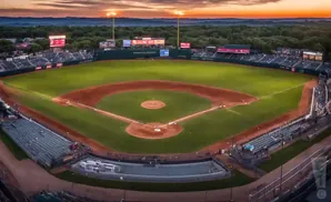 Aerial view of a baseball stadium at sunset with the field lit up and trees in the background.