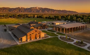 Aerial view of a ranch with wooden buildings, mountains, and fields at sunset.