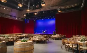 A venue set up for an event with tables and chairs, a stage with a blue backdrop, and red curtains.