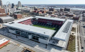 Aerial view of a modern stadium with red and black seats, a green field, and surrounding city buildings.