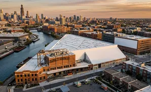 Aerial view of a triangular building with a white roof next to a river in an urban setting.