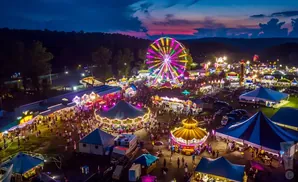 An aerial view of a brightly lit carnival at dusk with a Ferris wheel, carousels, and crowds of people.