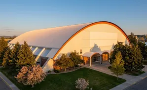 An aerial view of a large, white, arched building with an orange trim, surrounded by trees and green lawns at sunset.