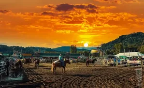 Rodeo arena at sunset with cowboys on horses and a vibrant orange sky.
