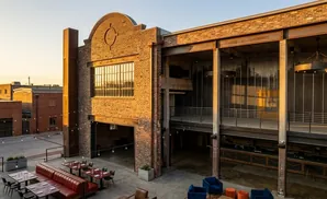An outdoor patio with tables and seating is adjacent to a modern brick building with large windows and industrial-style beams.