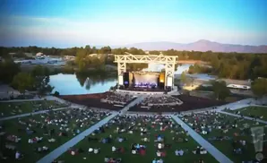 Outdoor concert stage with a large audience seated on a grassy lawn near a lake.