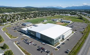 Aerial view of a modern indoor sports complex with large windows, surrounded by parking lots, sports fields, and residential areas.