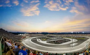 Spectators watch a race at a speedway under a sunset sky with clouds.