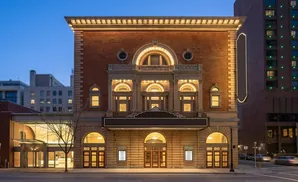 Ornate brick theater with arched windows and doors, lit up at dusk, next to a modern high-rise.