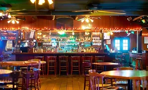 Interior of a dimly lit bar with wooden stools, tables, and a well-stocked liquor shelf illuminated by string lights.