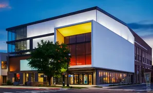 Modern building with glass facade, lit-up upper floors, and tree in front at dusk.