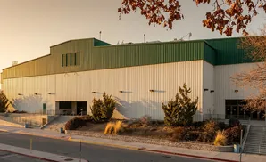 A large white and green building with sloped roofs, illuminated by warm evening light, with trees and a street in the foreground.