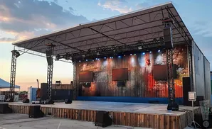 Outdoor stage setup with a corrugated metal backdrop, screens, speakers, and lighting.