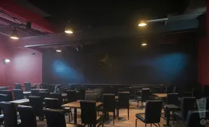 An empty, dimly lit room with rows of black chairs and tables, set up for a performance or presentation.