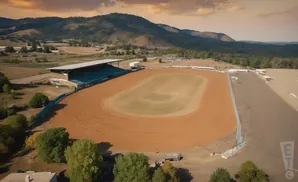 Aerial view of a dirt race track with grandstands, surrounded by trees and hills under a cloudy sky.